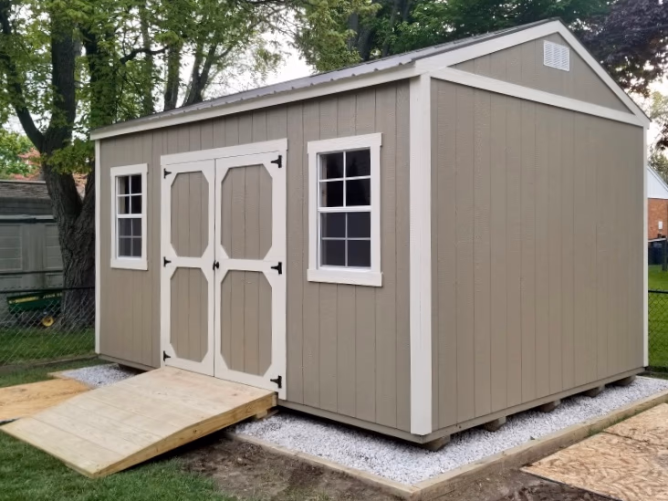 A tan garden shed on a white stone gravel pad with a wood ramp